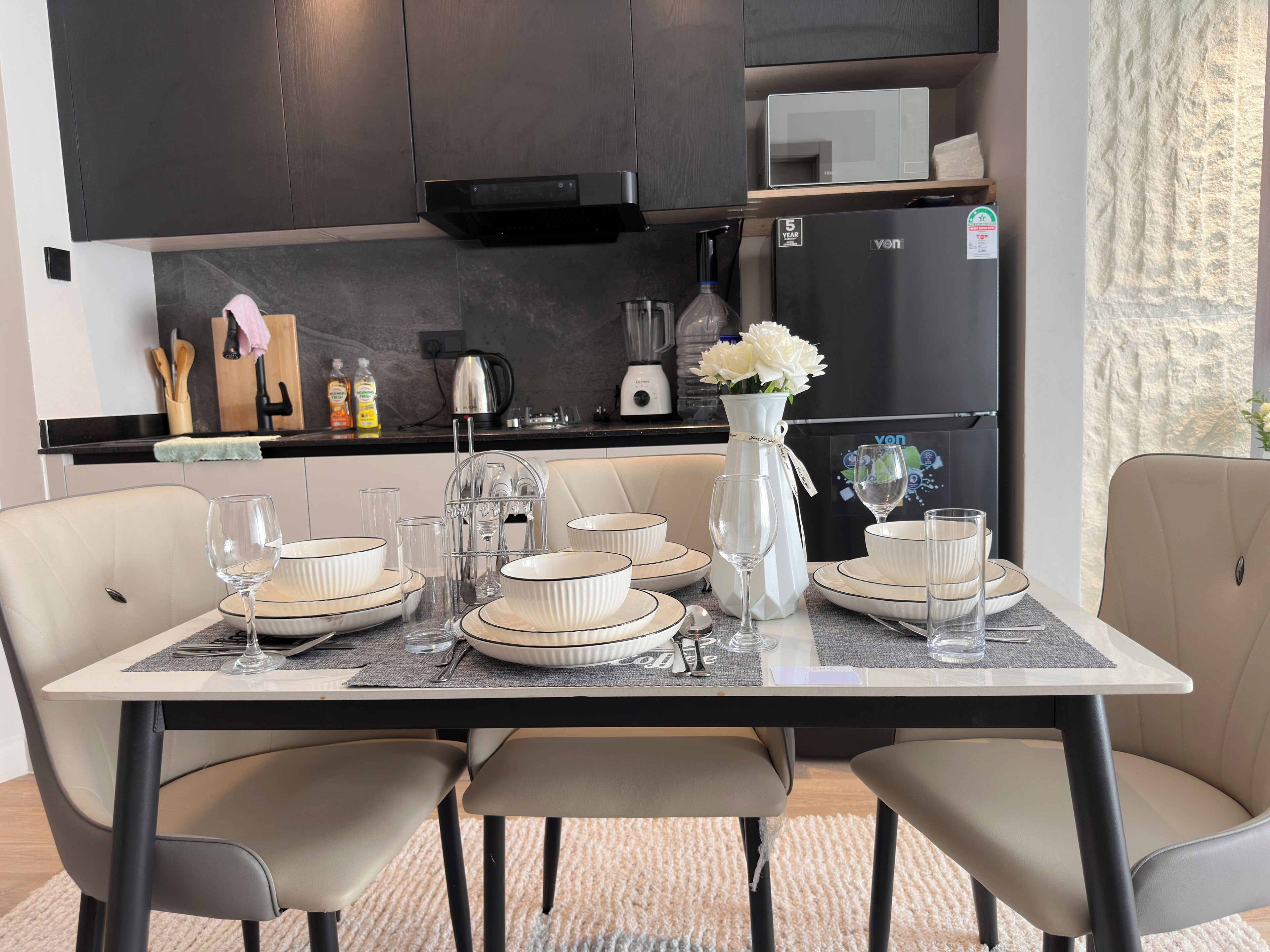 Une cuisine moderne avec des meubles de couleur sombre, un réfrigérateur et un coin repas agrémenté d’assiettes, de bols et de verres blancs.