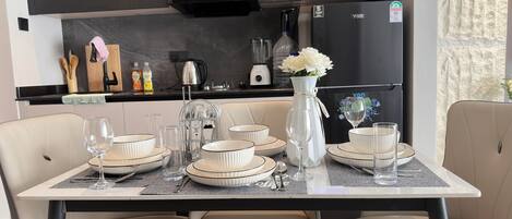 Une cuisine moderne avec des meubles de couleur sombre, un réfrigérateur et un coin repas agrémenté d’assiettes, de bols et de verres blancs.