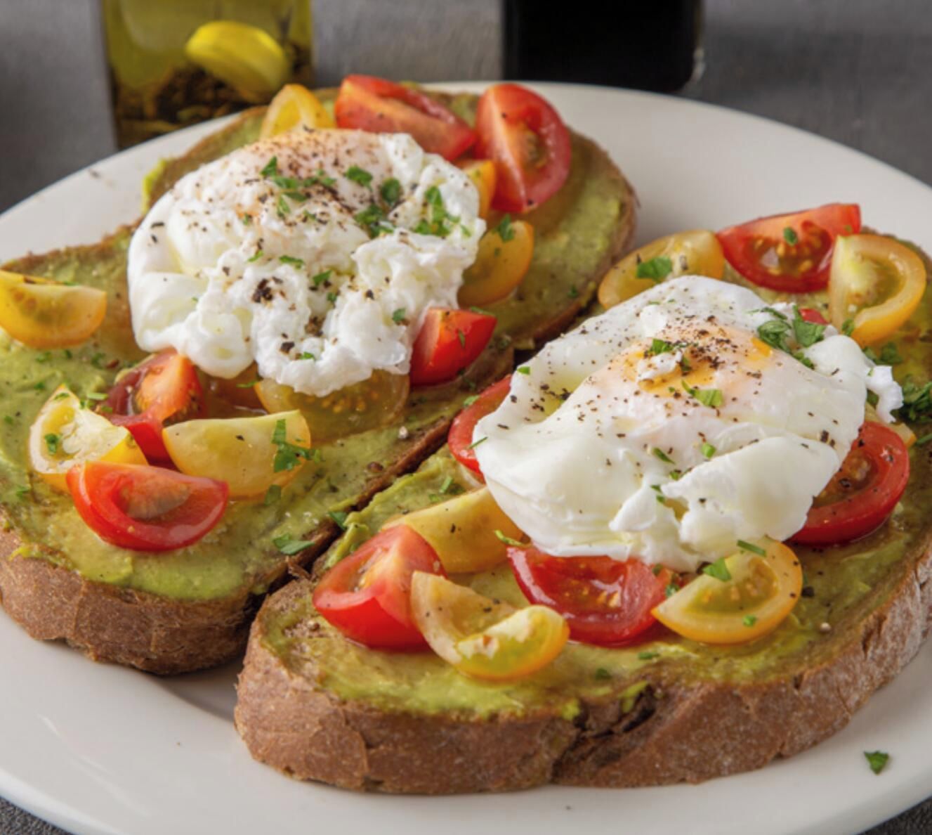 Een bord met avocado toast, belegd met gepocheerde eieren en plakjes tomaat.