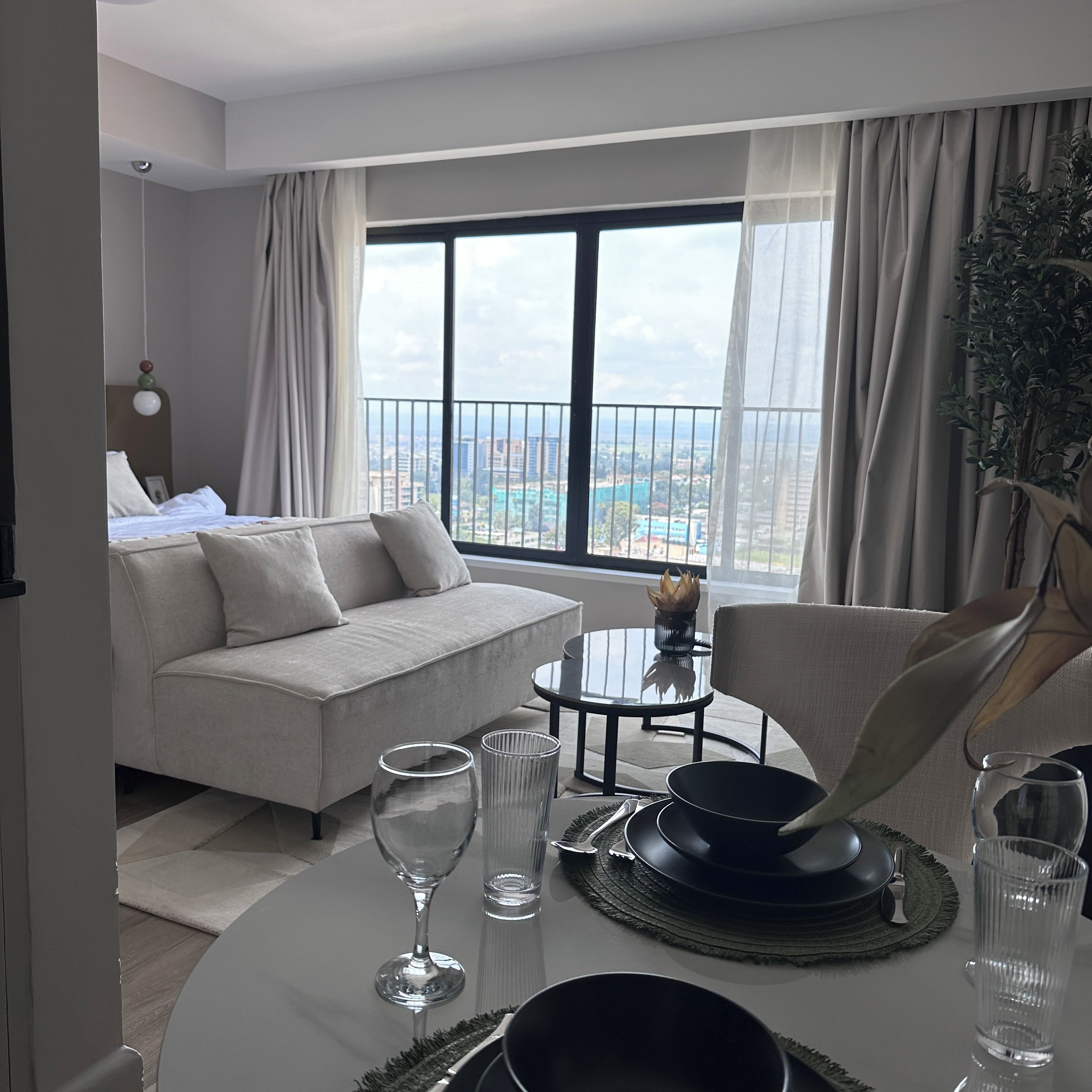 A modern hotel room with a balcony, a sofa, a dining table set for two, and a view of the cityscape.