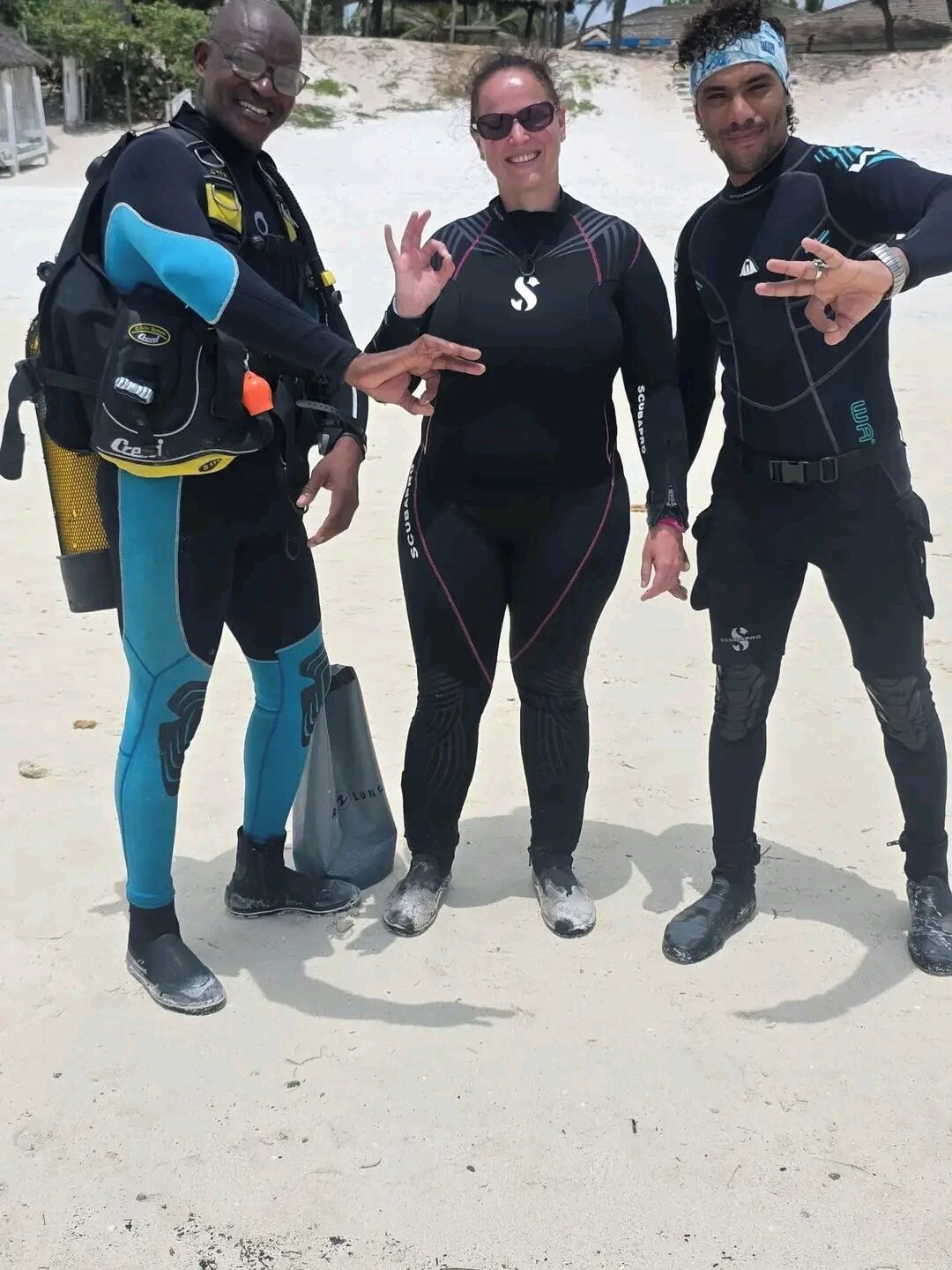 Three individuals in scuba diving gear standing on a sandy surface.