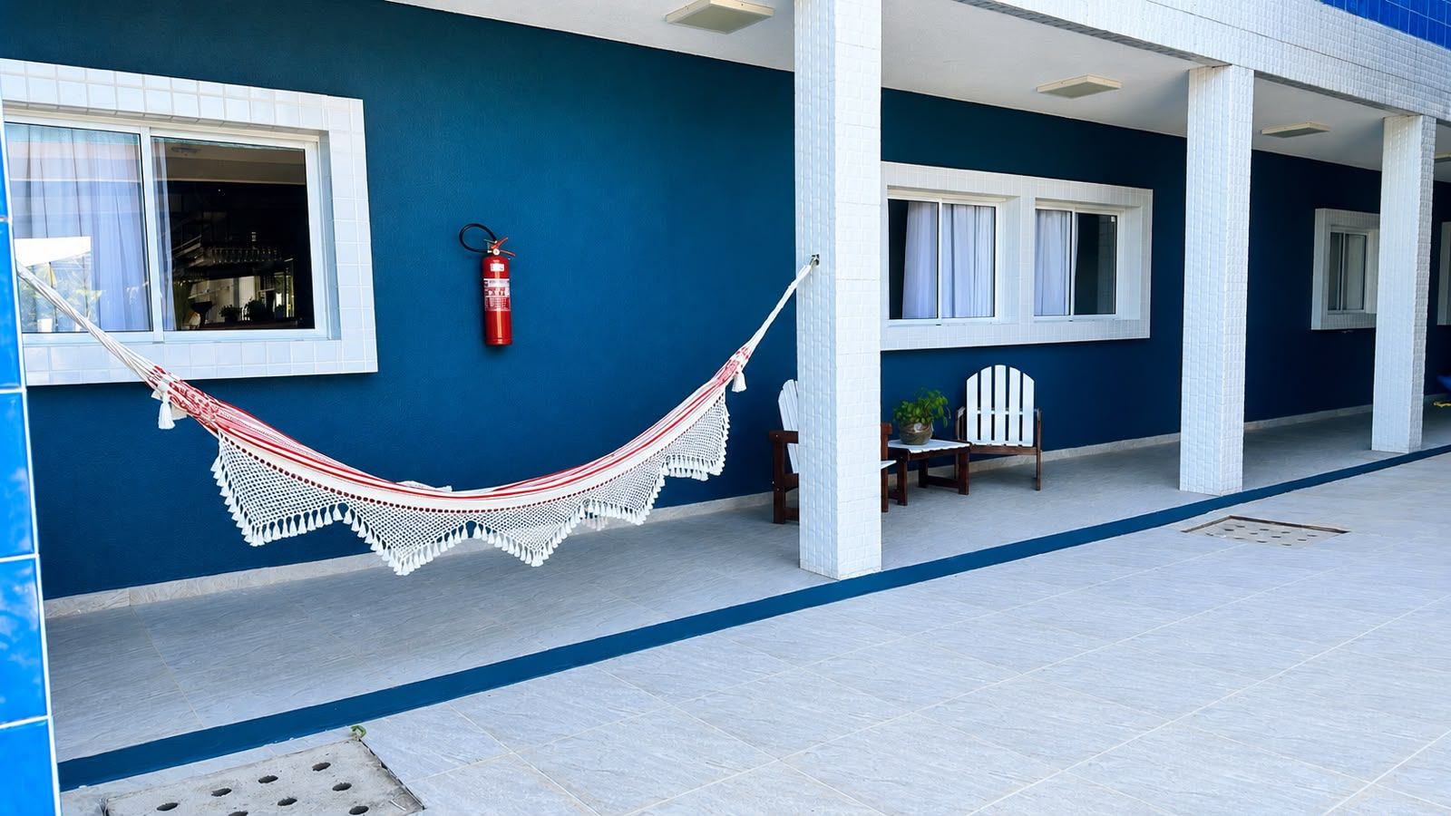 A blue exterior wall with a white tiled floor, a red fire extinguisher, a white hammock, and two wooden chairs.