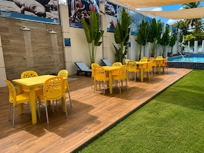 A poolside area with yellow tables and chairs, a wooden deck, and a grassy area.