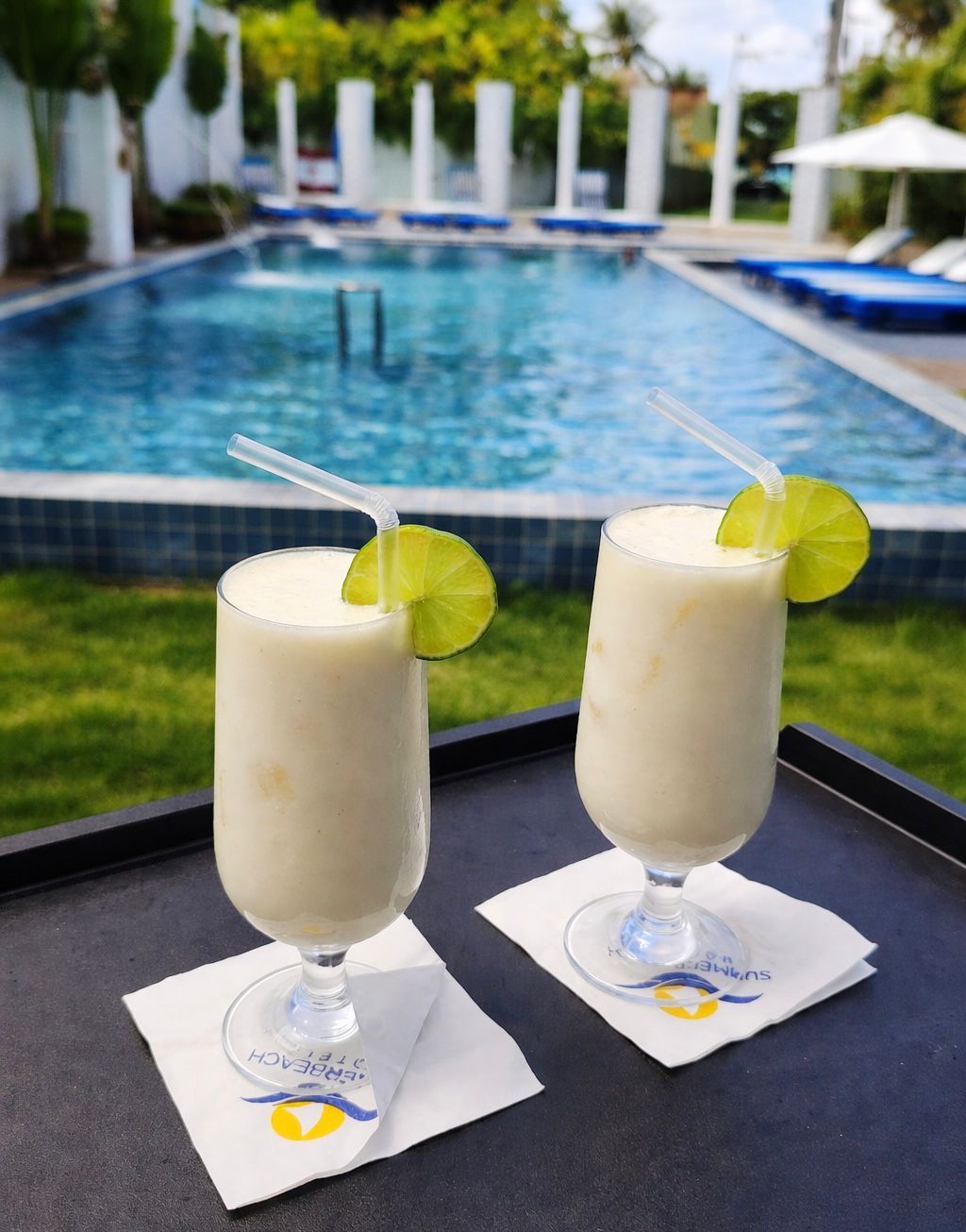 Two glasses of a beverage with lime garnish on a tray by a poolside.