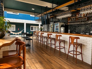 A bar area with wooden chairs, a counter with a TV, and a view of a blue building. - Hotel Summer Beach Paripueira Maceió (Paripueira)