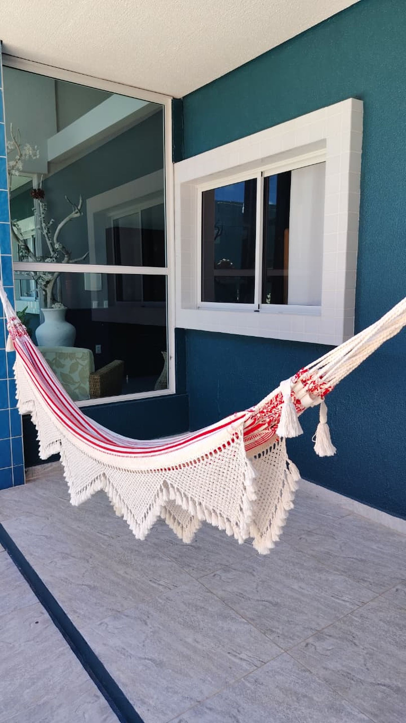 Una hamaca atada a un ventanal con marco blanco sobre un muro azul, con piso de baldosas y un área exterior visible.