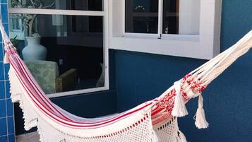 Una hamaca atada a un ventanal con marco blanco sobre un muro azul, con piso de baldosas y un área exterior visible.