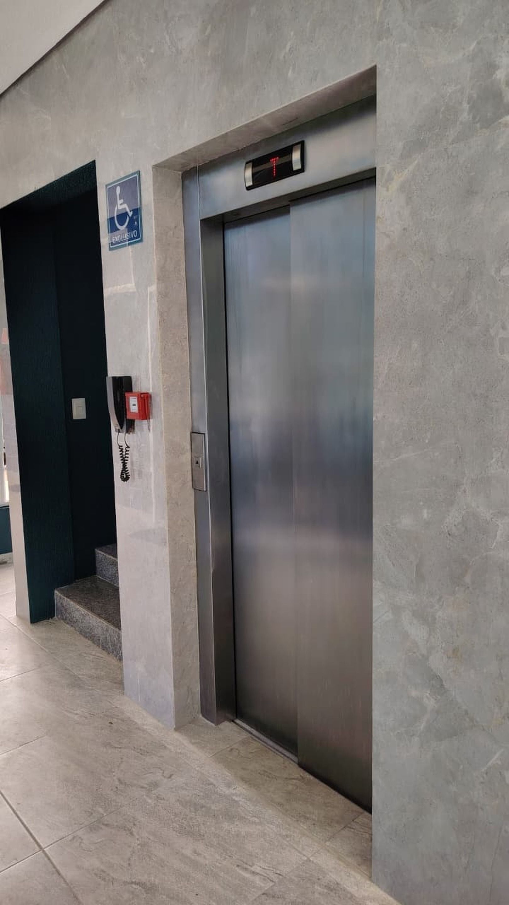 Un ascensor con pantalla digital, señal de accesibilidad para sillas de ruedas y un teléfono rojo de emergencia.