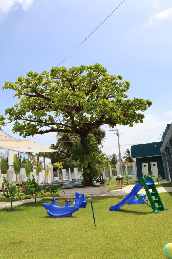 Un grand arbre se dresse dans un espace verdoyant, agrémenté d’un terrain de jeux avec notamment un toboggan et des balançoires.