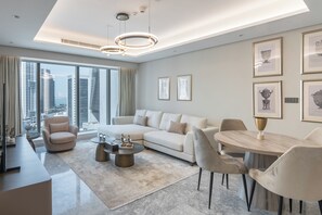 A modern living room with a sofa, chairs, a coffee table, and a dining table. There are framed pictures on the wall and a view of tall buildings through the window.