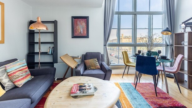 Ein modernes Wohnzimmer mit Sofa, Sessel, Couchtisch und Essbereich. Ein Bücherregal, ein Gemälde und ein großes Fenster mit Blick auf die Stadt sind ebenfalls zu sehen.