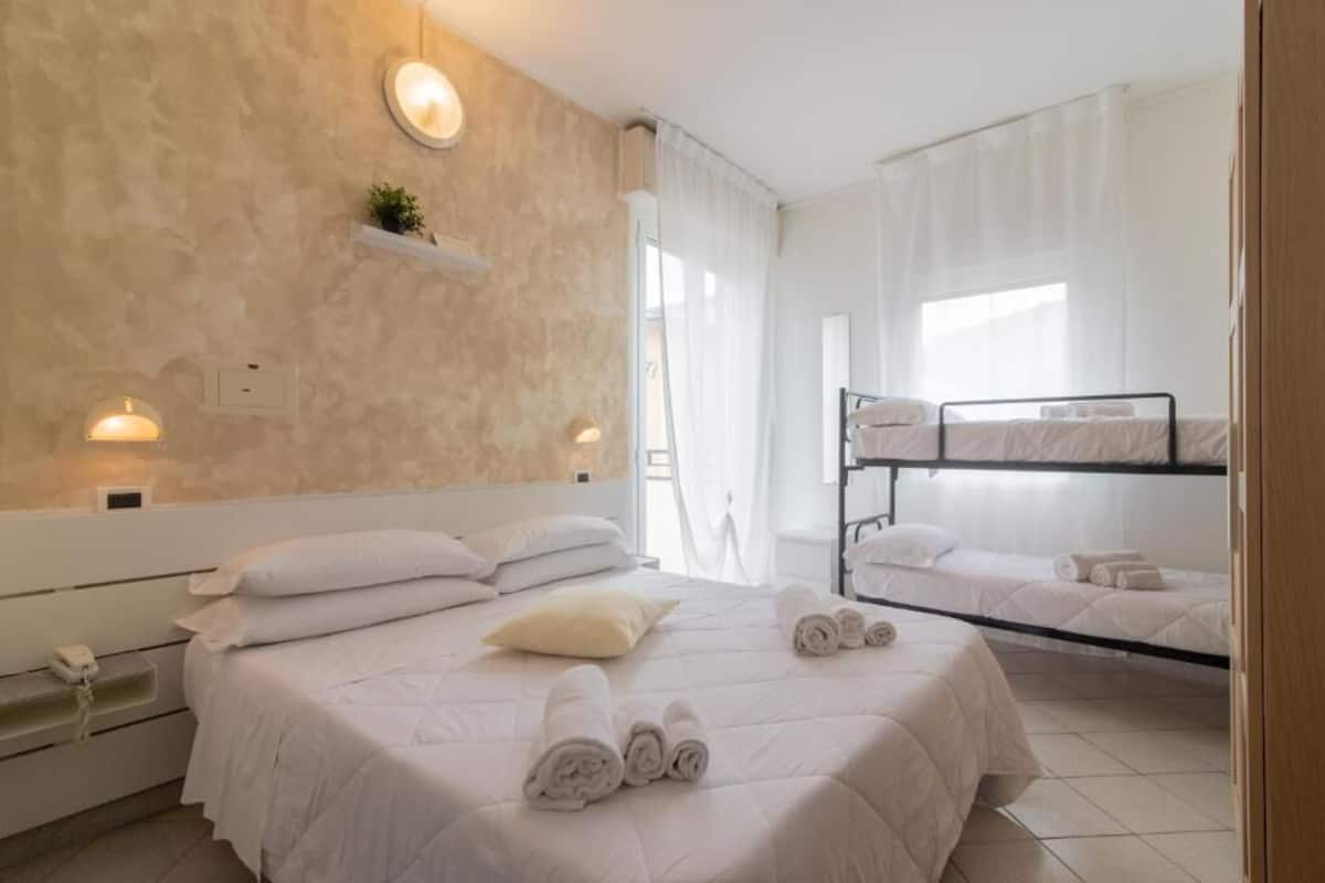 Una habitación de hotel con una cama grande, una litera, una mesita de noche con lámpara y un pequeño estante con una planta.