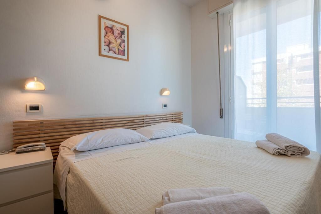 Et hotelværelse med seng, sengeborde med lamper, et billede på væggen og et vindue med gardiner.
