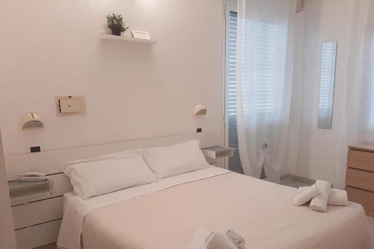 Habitación de hotel ordenada con cama, mesitas de noche, lámpara, estante pequeño y ventana con cortinas.