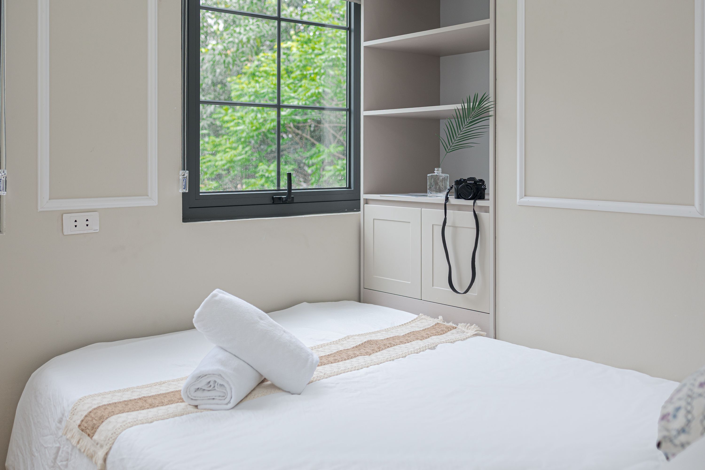 A neatly made bed with a rolled towel, a window with a view of greenery, and a camera on a shelf.