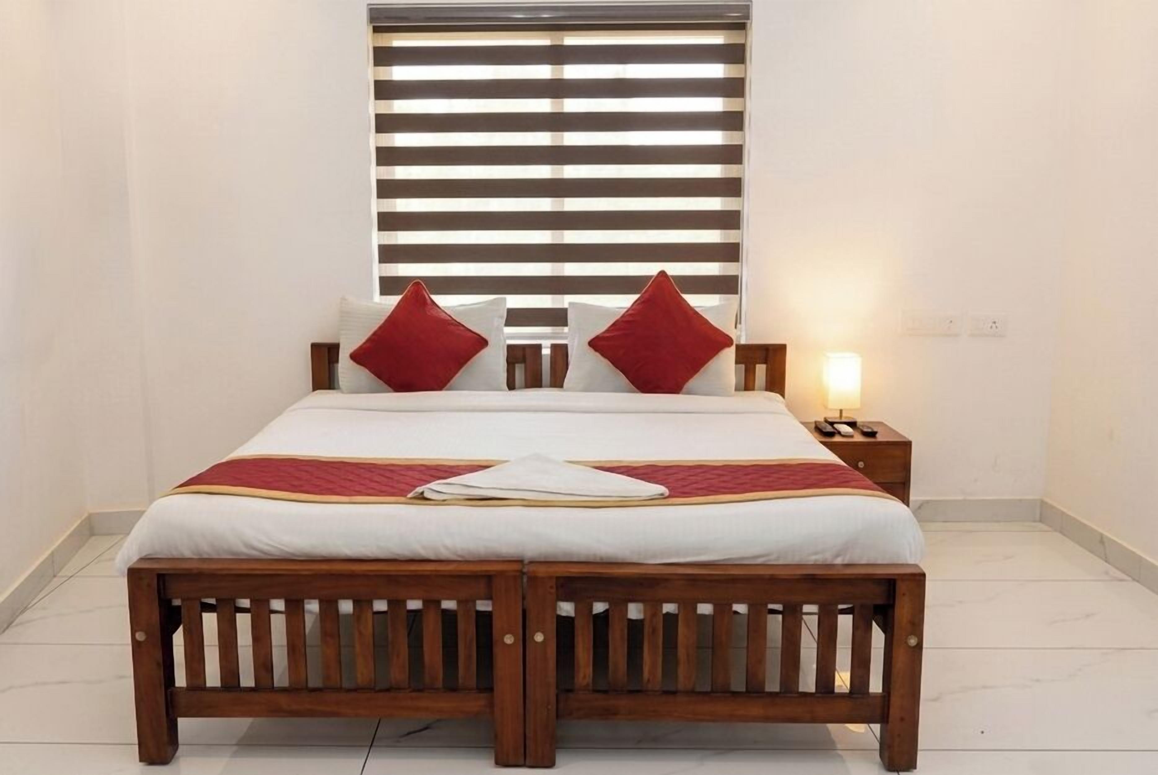 A bed with a wooden frame, white bedding, red pillows, a bedside lamp, and a nightstand with a phone.