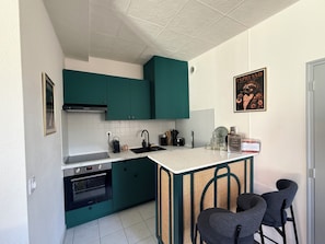 Une cuisine compacte avec des meubles de couleur turquoise, un plan de travail blanc, un four encastré, un évier et deux tabourets de bar.