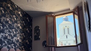 A bedroom with a bed, a window with a view of a building, a wall with a floral pattern, and a ceiling fan. - The Zebra Club (Saumur)