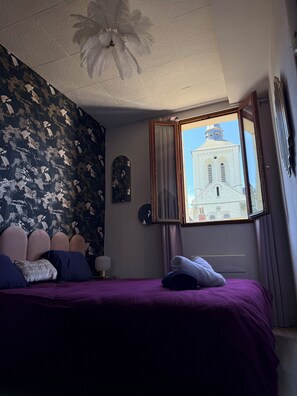 Une chambre à coucher avec un lit, une fenêtre donnant sur un immeuble, un mur orné d’un motif floral et un ventilateur de plafond.