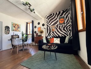 Un salon avec un mur orné d’un motif zébré, une table basse ronde, un canapé, un coin repas avec des chaises et une radio ancienne.