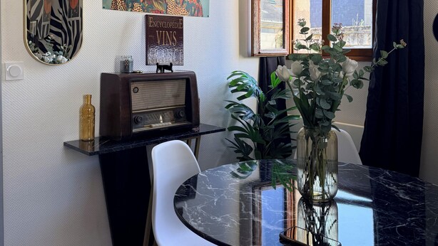 A dining area with a glass table, white chairs, a vintage radio, a painting, and a window with curtains. - The Zebra Club (Saumur)