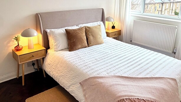 Ein ordentlich eingerichtetes Schlafzimmer mit einem großen Bett, zwei Nachttischen mit Lampen, einem Fenster mit Vorhängen und einem Heizkörper.