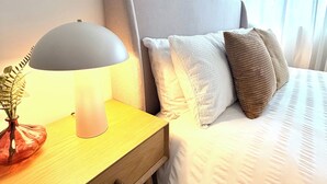 Ein ordentlich bezogenes Bett mit grauem Kopfteil, ein Nachttisch mit Lampe und eine Pflanze in einer Vase.
