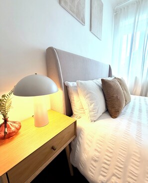 Ein ordentlich bezogenes Bett mit grauem Kopfteil, ein Nachttisch mit Lampe und eine Pflanze in einer Vase.