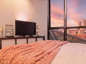 Ein Schlafzimmer mit einem Bett, einem Fernseher auf einem Schrank, einem Fenster mit Blick auf die Stadt und einem dekorativen Gegenstand auf dem Schrank.