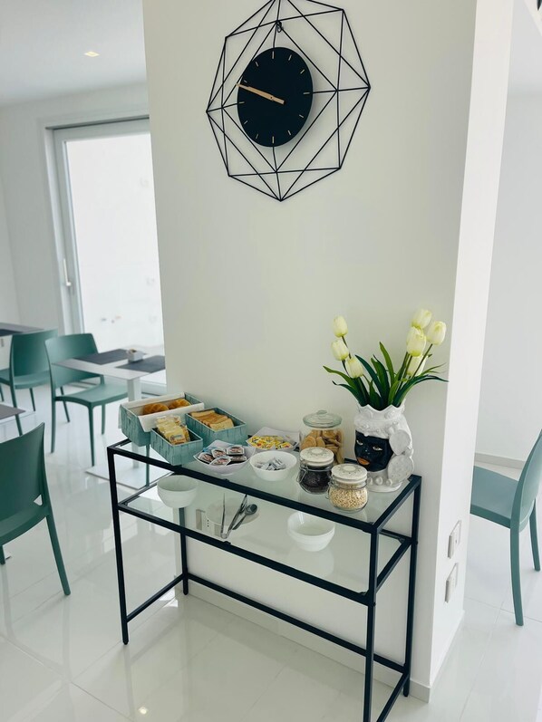 A modern dining area with a geometric clock, a buffet table with food items, and a vase with white flowers. - Gheloi Apartments (Gela)
