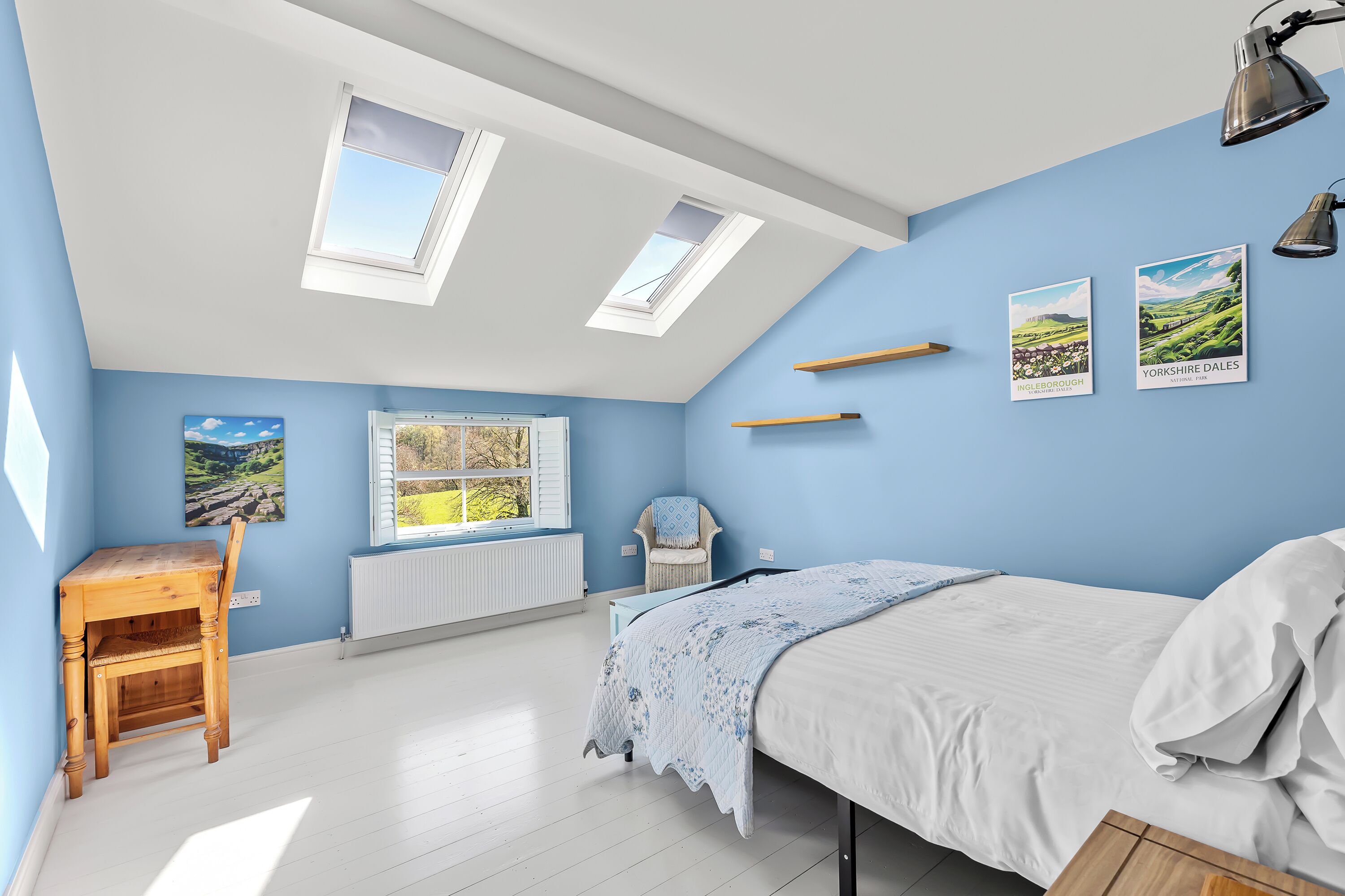 Une chambre avec un puits de lumière, un lit, un bureau, une chaise et deux affiches représentant des paysages accrochées au mur.