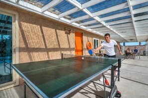 Ein Tischtennis-Tisch steht draußen auf einer Terrasse, die von einer Glasüberdachung überspannt ist.
