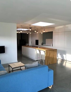 Ein modernes Wohnzimmer mit einem blauen Sofa, einem kleinen Couchtisch und einer Küchenzeile mit einer Holzinsel und weißen Barhocker.