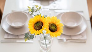 Ein Tisch ist mit weißen Tellern, Schüsseln und Besteck gedeckt, in der Mitte steht eine Vase mit Sonnenblumen.