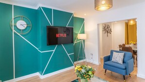 Ein modernes Wohnzimmer mit türkisfarbenen Wänden, einem an der Wand befestigten Fernseher, einem blauen Sofa, einer Stehlampe und einem Essbereich im Hintergrund.