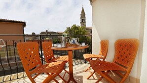 Ein Balkon mit orangefarbenen Stühlen und einem Tisch, einer Uhr sowie Blick auf einen Turm.