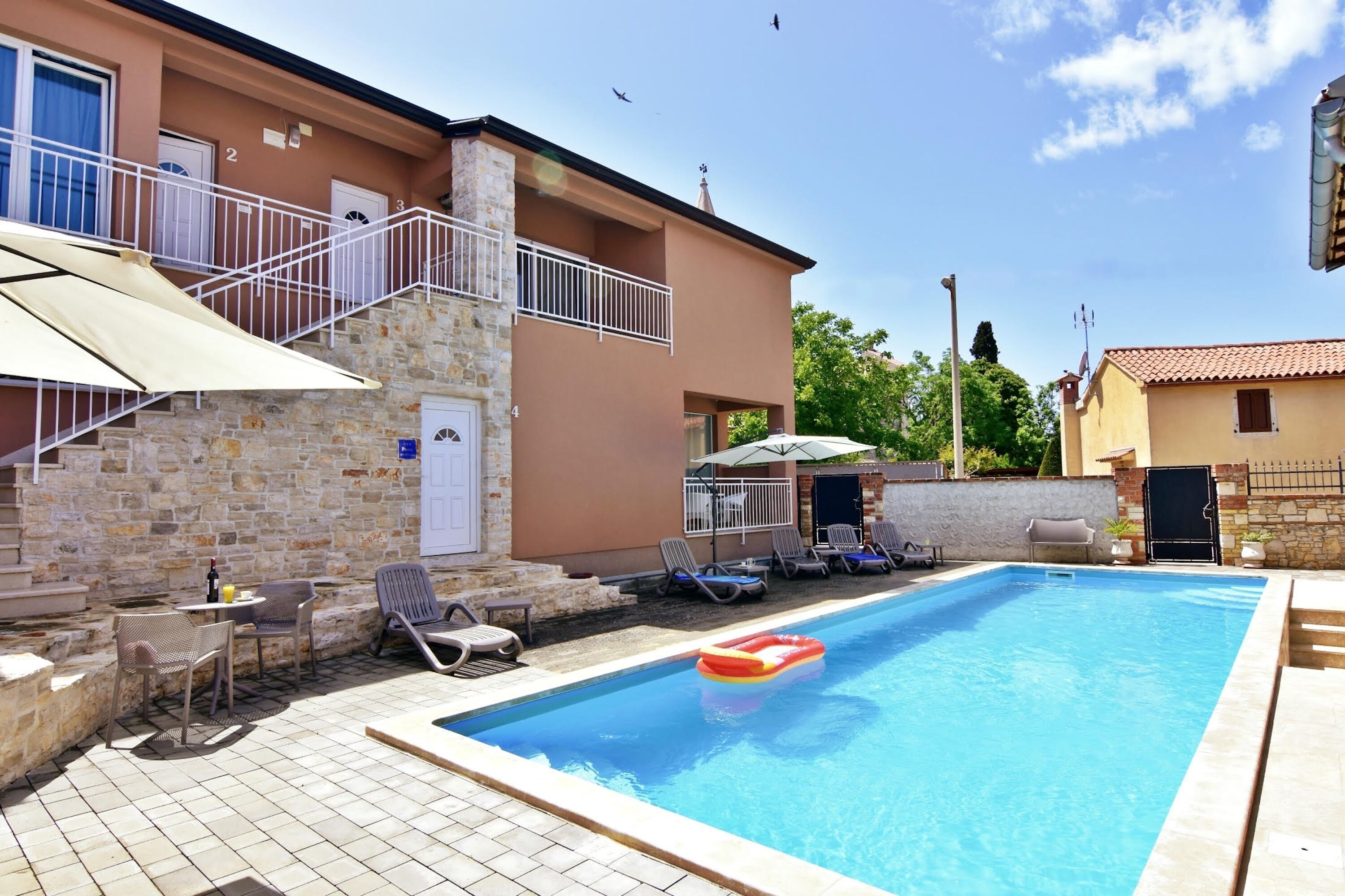 A swimming pool with lounge chairs, a stone wall, and a staircase leading to a balcony.