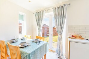 Un comedor con una mesa dispuesta para cuatro personas, sillas, una ventana con cortinas y una encimera de cocina con pan.