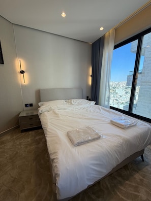A modern hotel room with a large bed, a nightstand, a wall-mounted light, and a view of the city through a large window. - 204 Tara-jre Al Lweibdeh 2bdr With Stunning View (Amman)