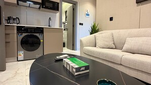 Uma sala de estar moderna com uma mesa de centro preta, um sofá bege e uma área de cozinha com eletrodomésticos.