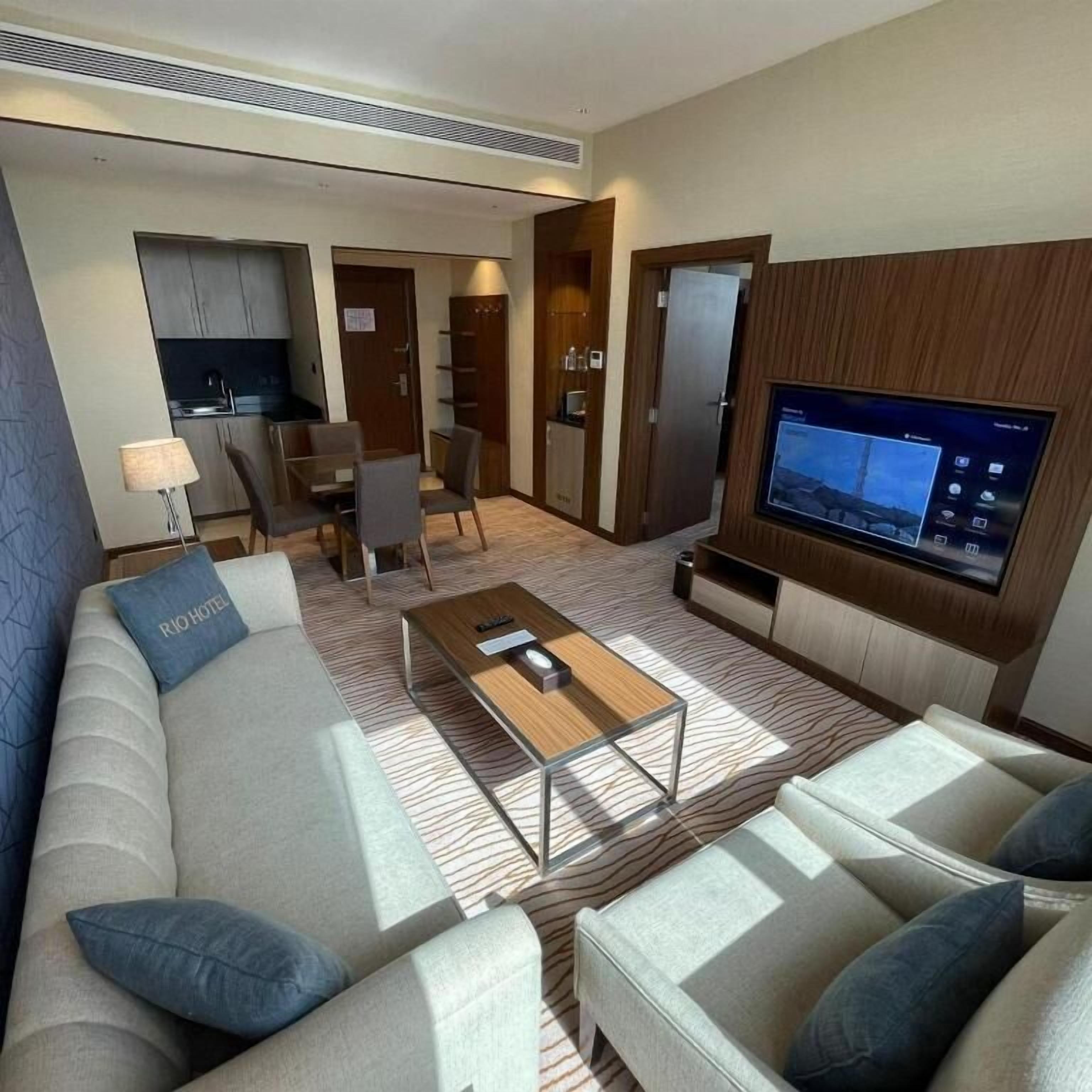 A hotel room with a sofa, a television, a coffee table, a dining area with chairs, and a kitchen area.
