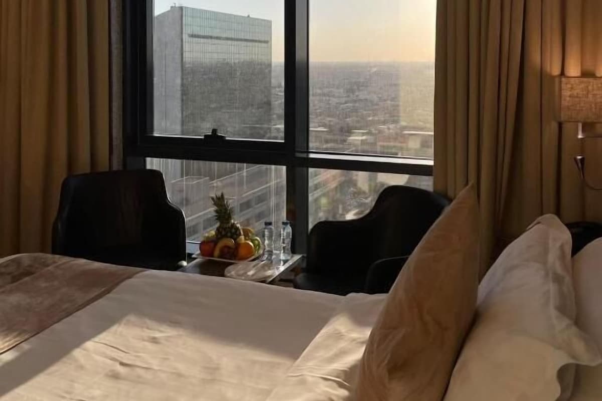 A hotel room with a large window, a bed, a table with fruit, and two chairs.