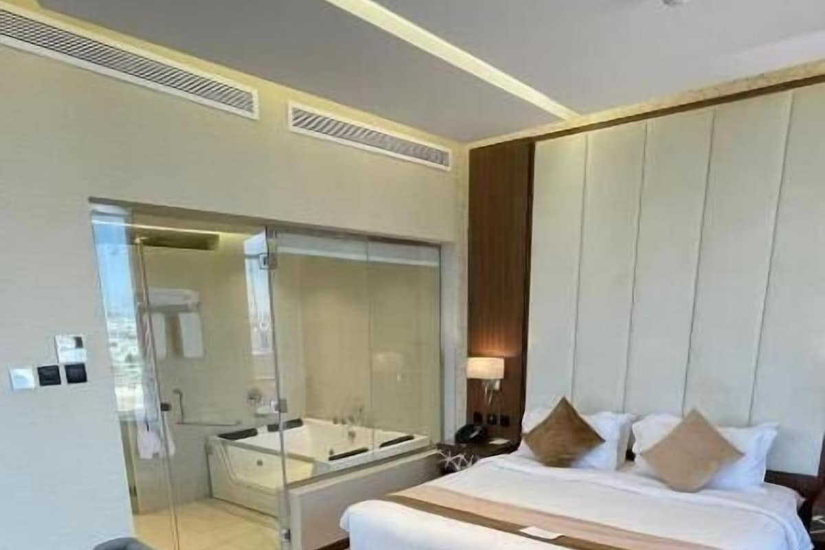 A hotel room with a large bed, a bedside table, a chair, a bench, and a glass-enclosed bathroom.