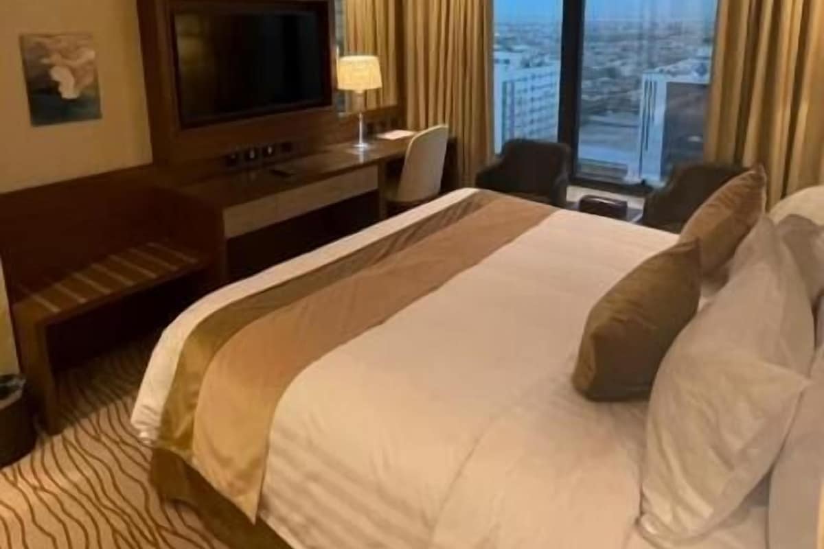 A hotel room with a large bed, a television, a desk with a lamp, and a view of the city.