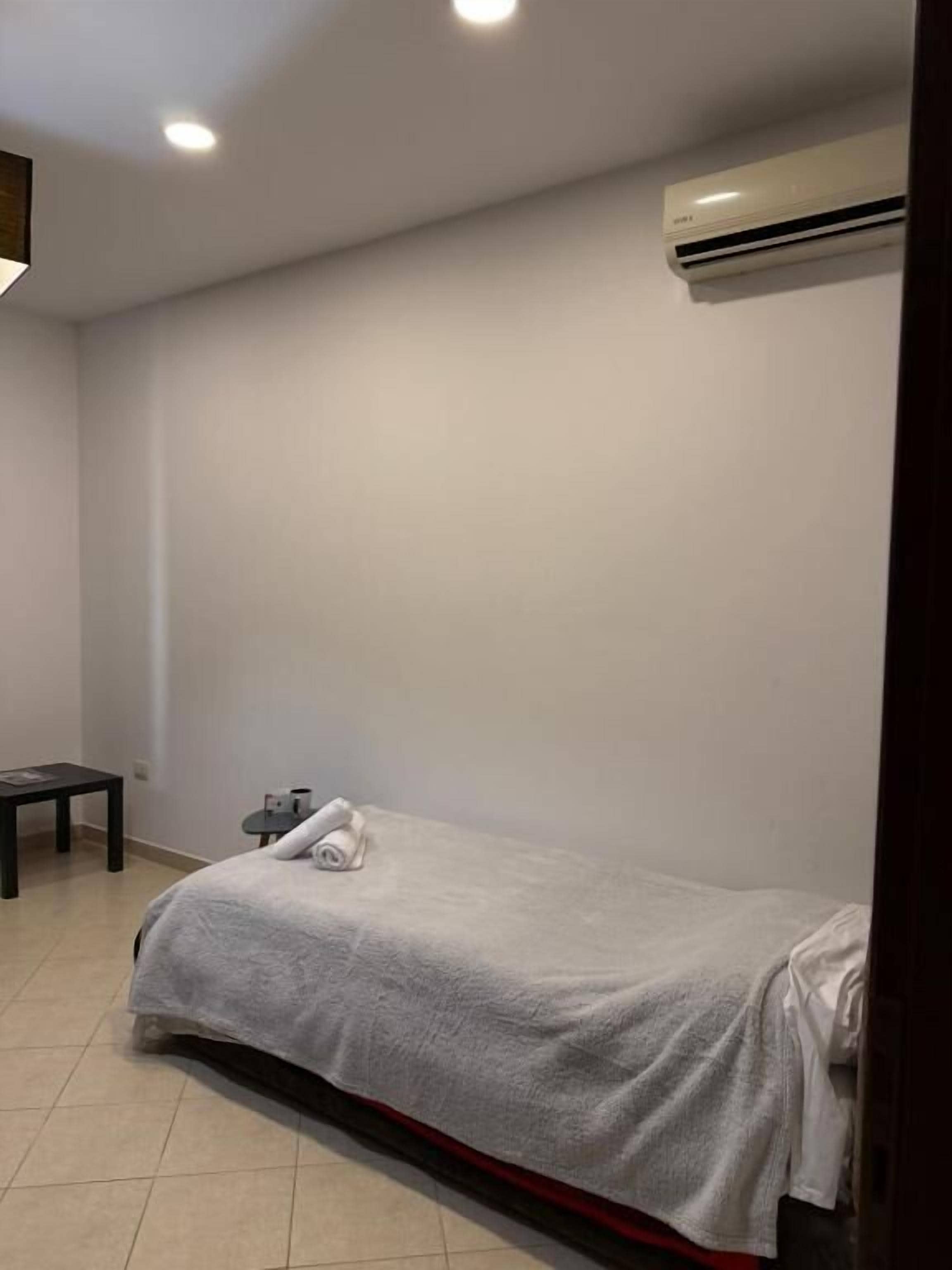A bedroom with a bed, a small table, an air conditioner, and a wall-mounted light fixture.