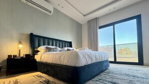 A hotel room with a large bed, a nightstand, a lamp, a view of the city through a large window, and an air conditioning unit. - Khozama Living (Jeddah)