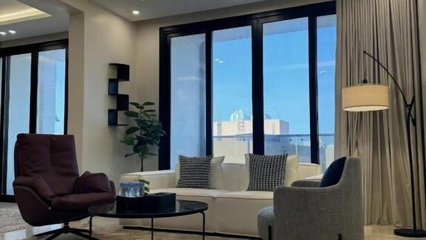 A modern living room with a sofa, two armchairs, a coffee table, and a floor lamp. Large windows offer a view of the cityscape. - Khozama Living (Jeddah)