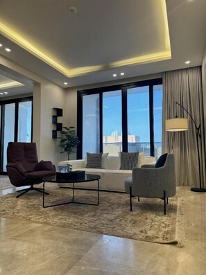 A modern living room with a sofa, two armchairs, a coffee table, and a floor lamp. Large windows offer a view of the cityscape. - Khozama Living (Jeddah)