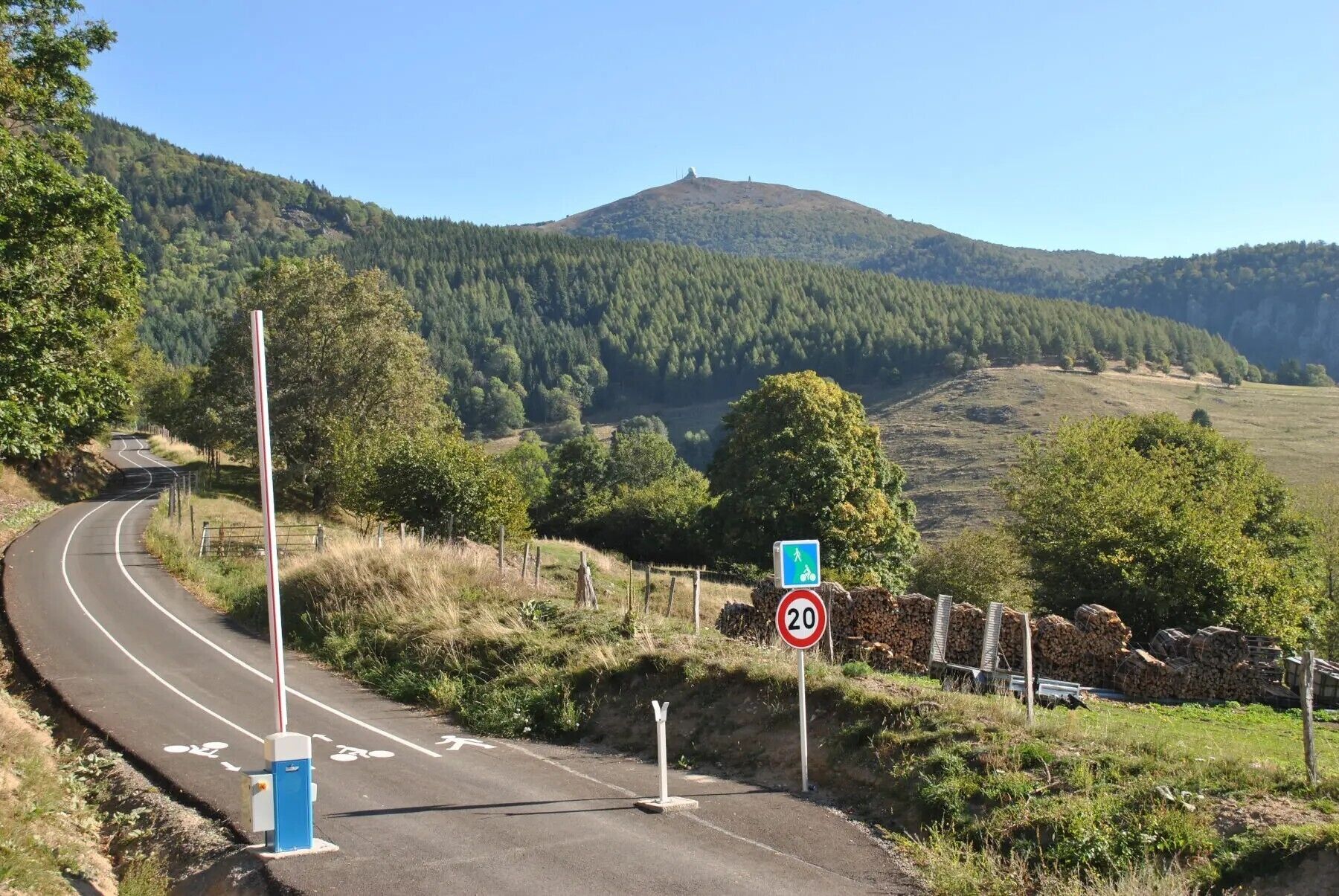 Une route sinueuse, avec un panneau indiquant la vitesse maximale autorisée, un panneau signalant un chemin piétonnier et une piste cyclable, et une montagne en arrière-plan.