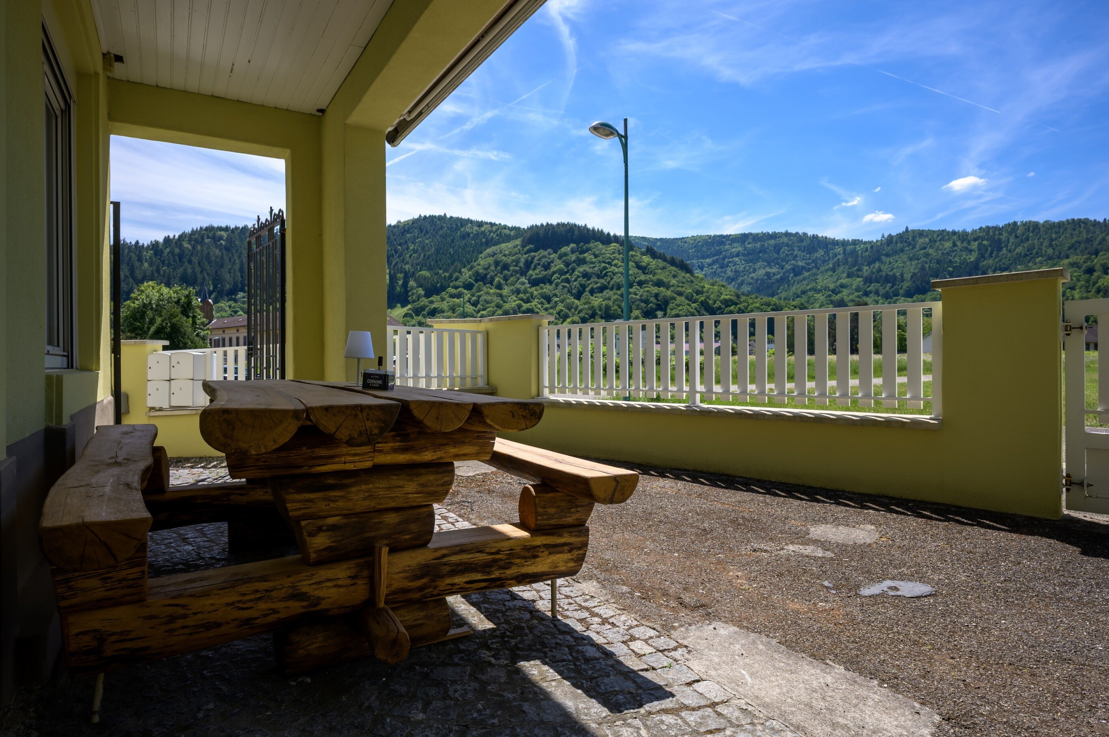 Une table et des bancs de pique-nique en bois, placés sous une marquise jaune, avec vue sur des collines verdoyantes et une clôture blanche.